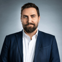 Bearded man with blue eyes wearing a white shirt and dark blazer against a gray background.