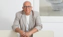 Bald man with glasses wearing a striped blazer over white shirt sitting against white wall with artwork.