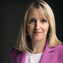 Blonde woman with blue eyes wearing a purple blazer and white shirt against a dark background.